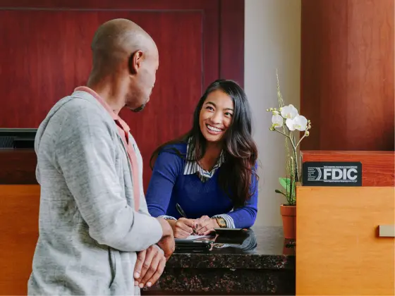 Bank employee assisting a customer at a counter with an FDIC sign and potted orchid.