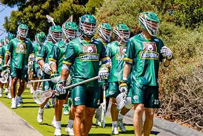 California Redwoods lacrosse players wearing green uniforms and helmets walk together outdoors on a path, holding their lacrosse sticks. Trees and bushes are visible in the background under a bright sky..