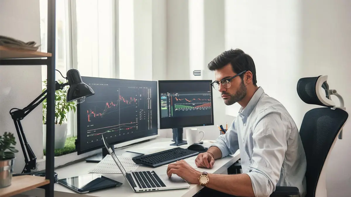 Photo of beared young man working intently on multiple computer screens