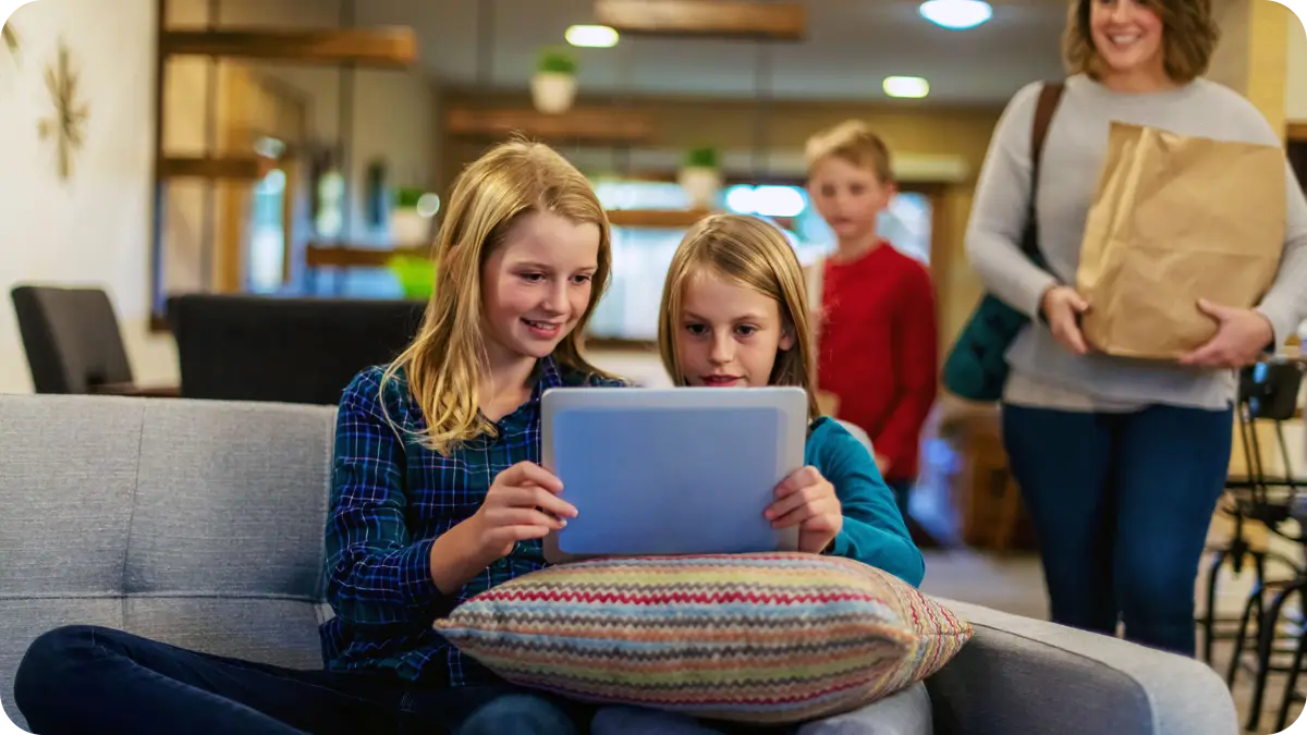 Two children look at a tablet while a woman holds a grocery bag behind them.