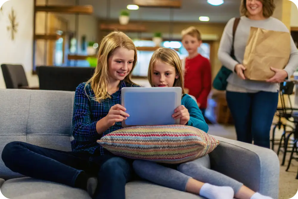 Children looking at a tablet as a woman holding a grocery bag stands in the background.