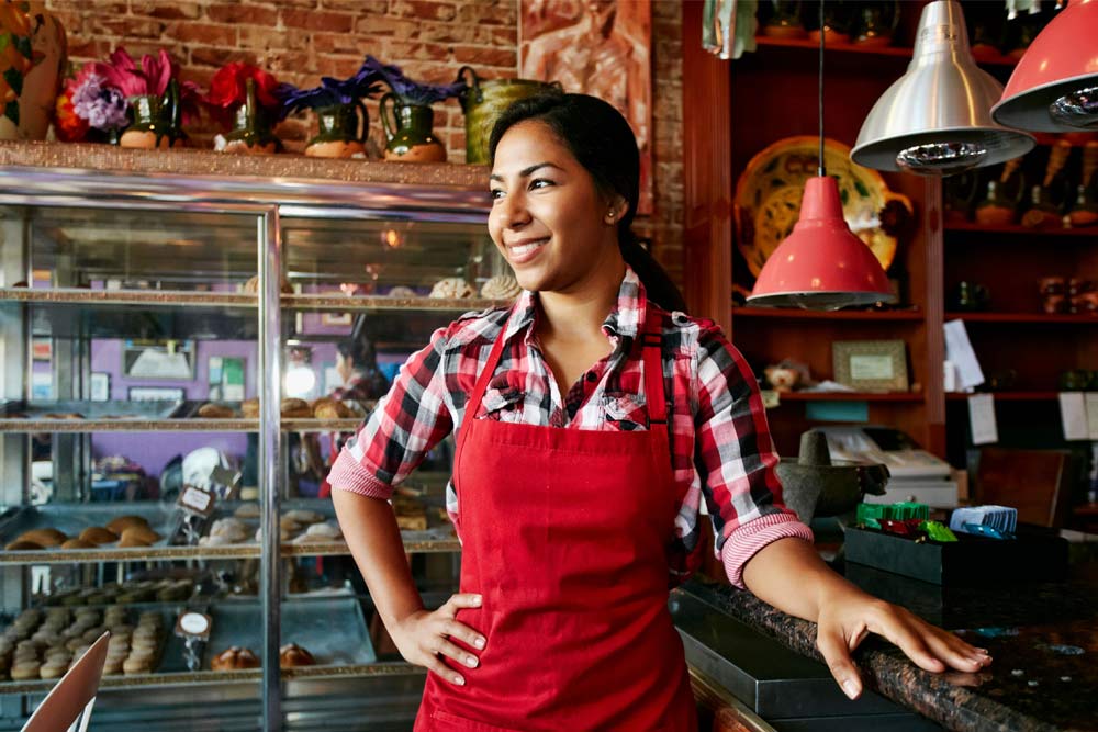 A woman wearing an apron in a bakery with baked goods in cases behind her.