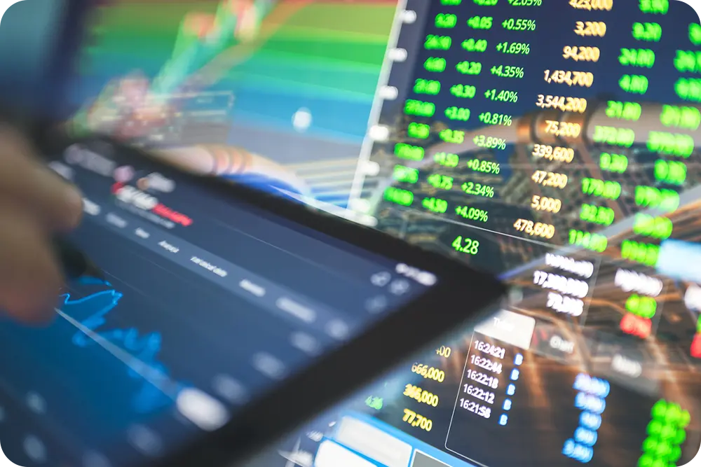 A hand holding a tablet in front of a computer monitor with data on it.