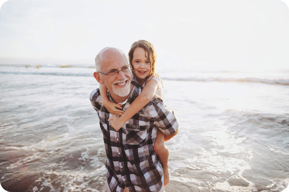 A man standing at the beach with a child on his back.