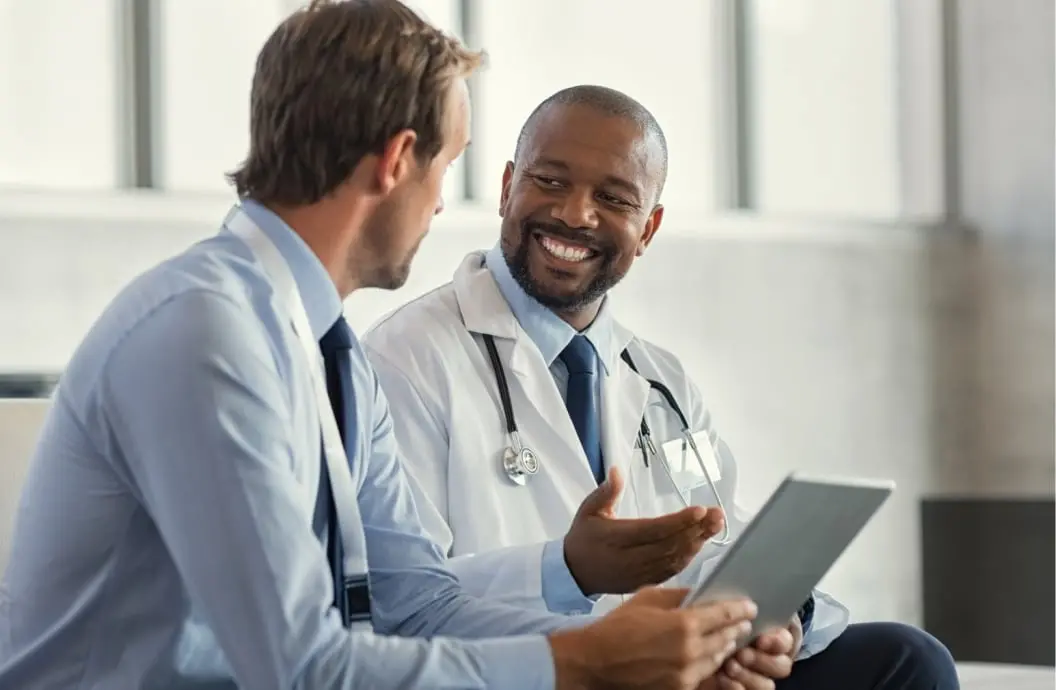 Doctor and a healthcare administrator sitting next to each other. The doctor has a stethoscope and the administrator is holding a tablet device.