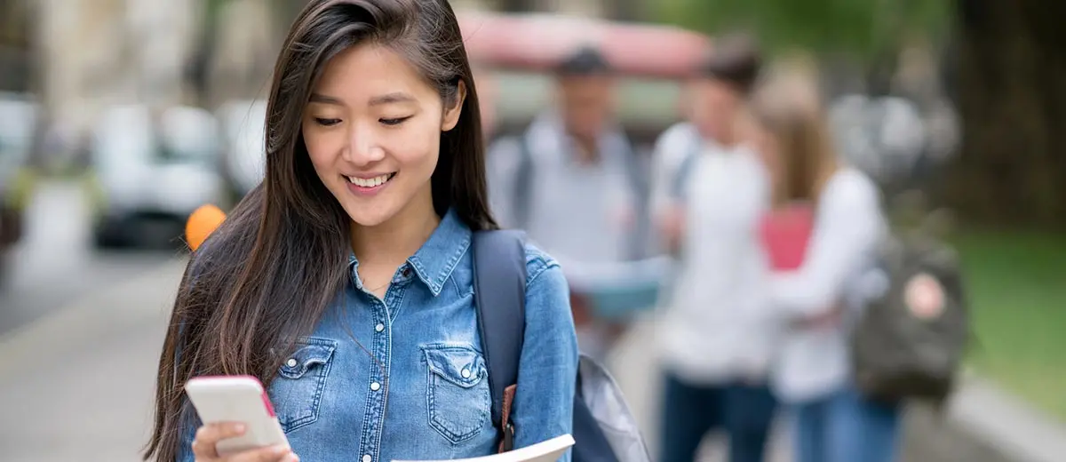 College-aged woman wearing a backpack and looking at her phone while walking on campus.