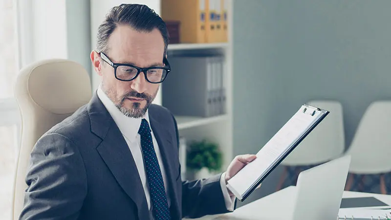 Man in a suit holding a clipboard while at his desk