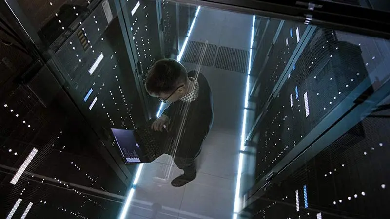 Overhead view of a man standing in a room with servers and looking at his laptop