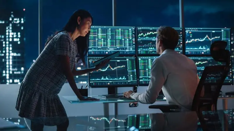 Man and woman in the office at night looking at six computer monitors and conducting an international ACH transaction