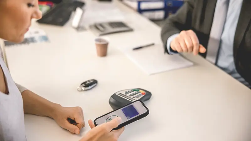 Person using contactless secure payment systems with a smartphone at a card reader while another person sits across the desk