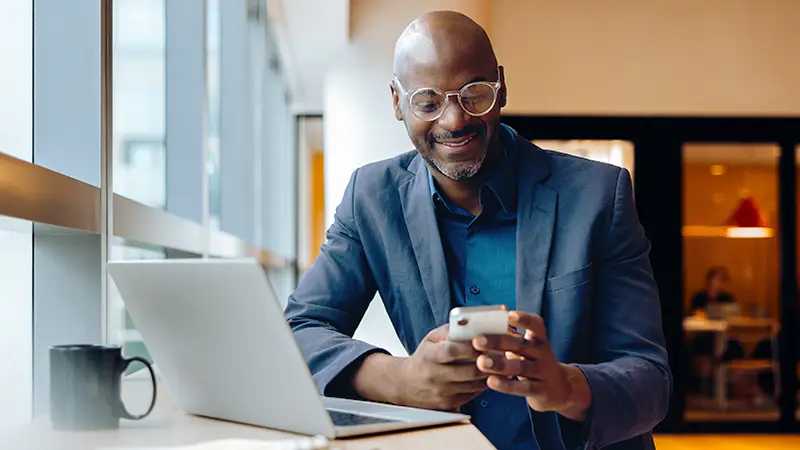 Professional using a smartphone while working on a laptop at a desk in an office setting.