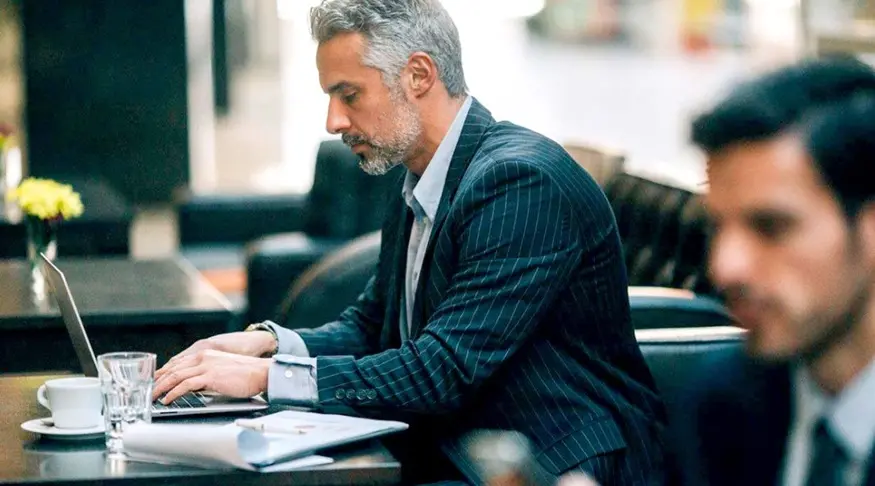 Businessman sits at a table researching the benefits of bank service providers versus boutique service providers on his laptop computer.