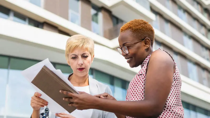Two women standing outside an office building and looking at paperwork about escheatment