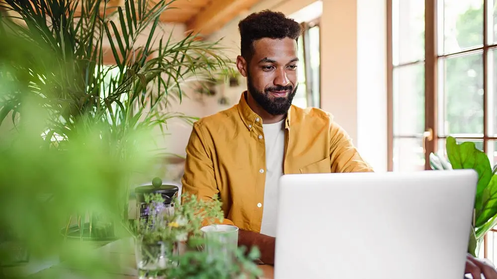 man in a yellow shirt surrounded by house plants and working on a computer