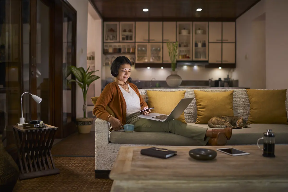 A woman sitting on a couch with a laptop and coffee cup, smiling while using a smartphone.  