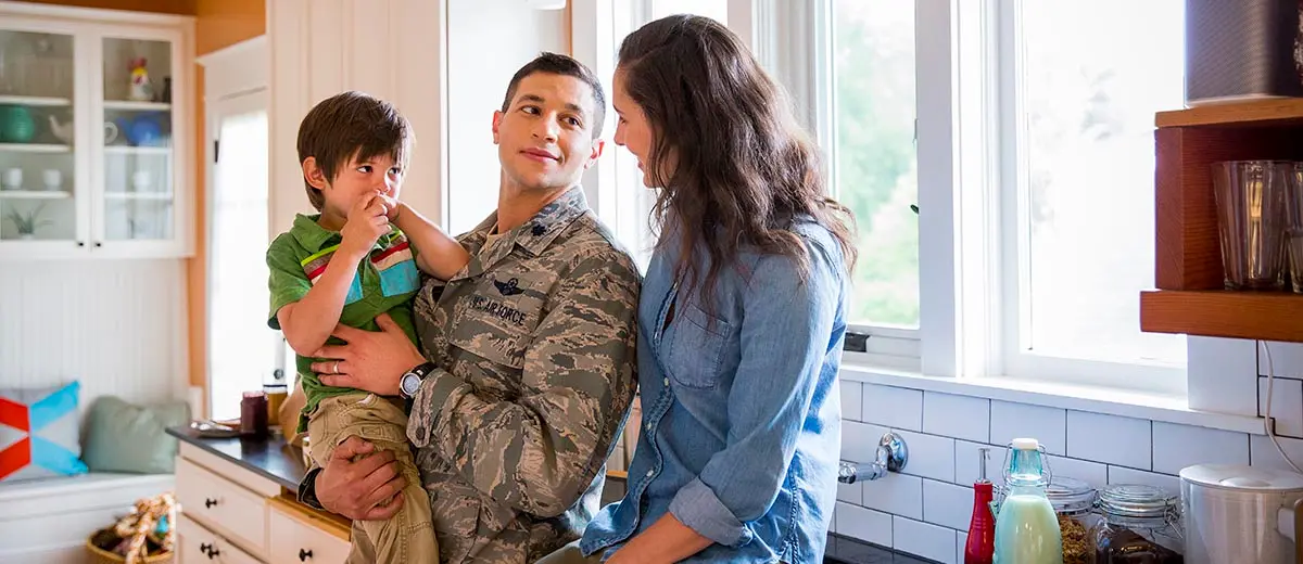 A military father holds his young son in the kitchen, while a woman smiles at them. 