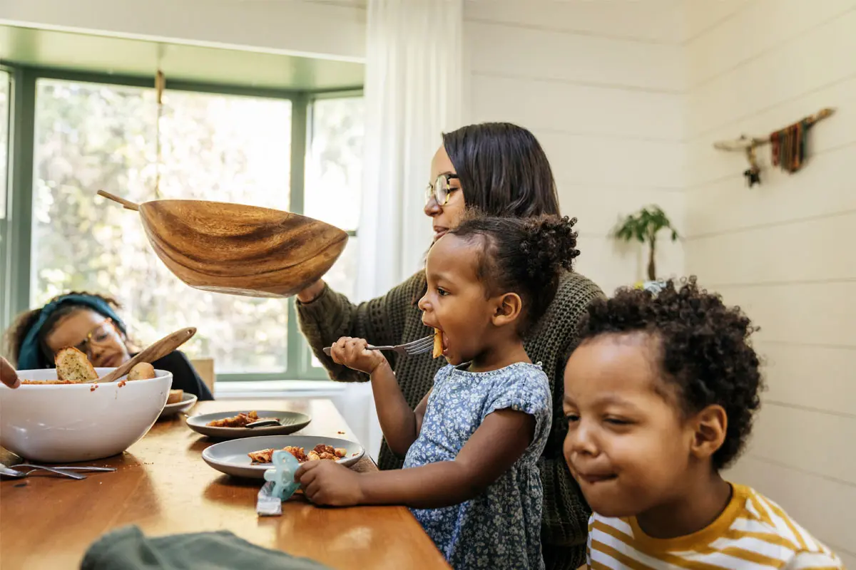 A woman serves food while two children eat at a wooden table in a sunny kitchen. 