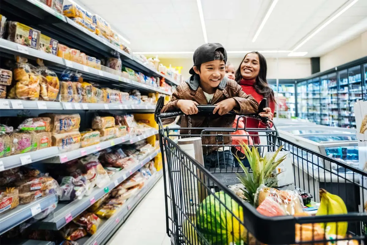 A child in a shopping cart smiles as they shop with a woman in a grocery aisle.