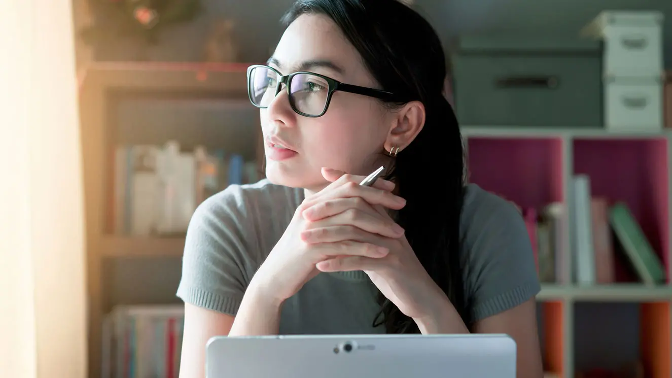 A woman with glasses sits at a desk, holding a pen and looking thoughtfully out a window. 