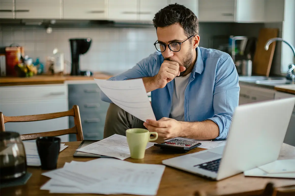 man in a blue shirt and glasses reading a piece of paper at his kitchen table