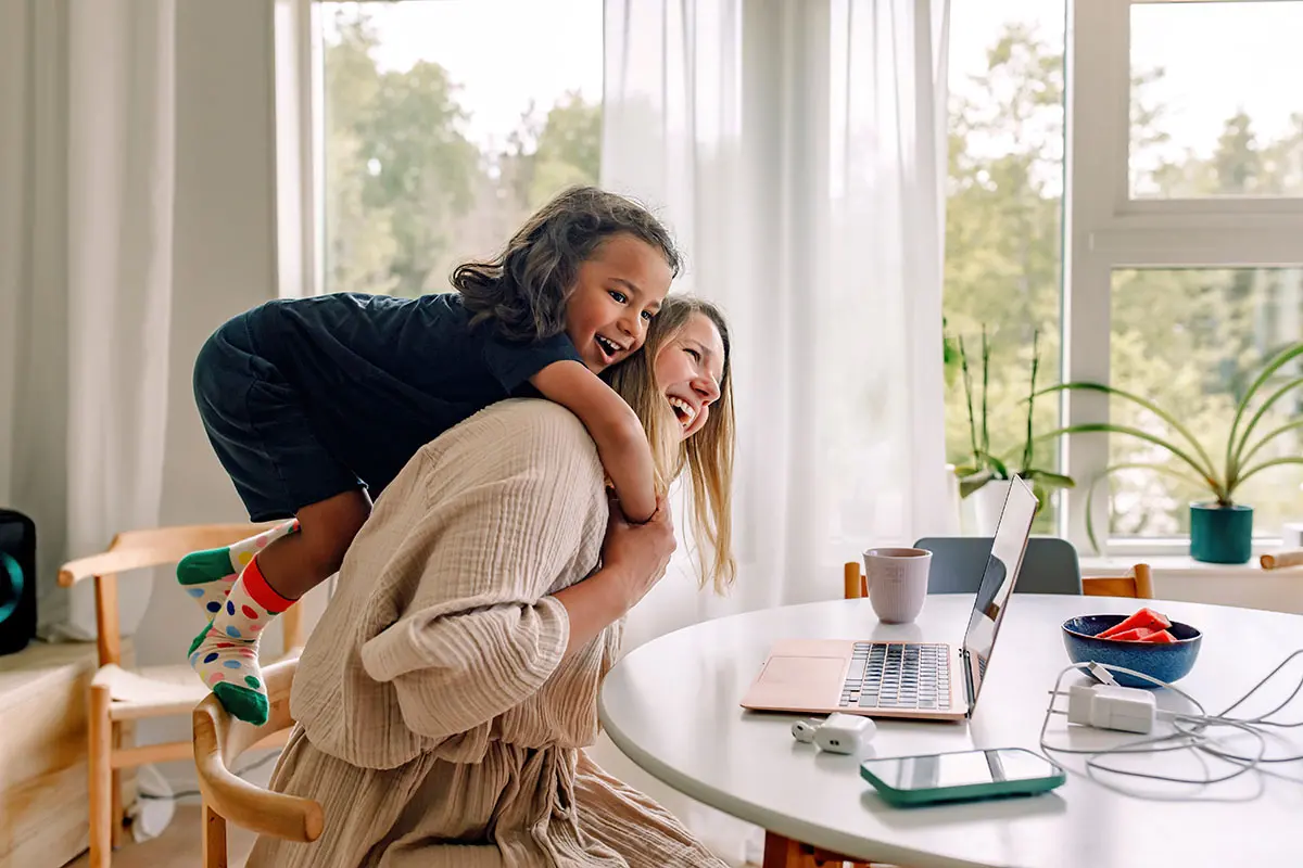 Woman working at a table with a laptop while a child climbs on their back in a sunlit room.