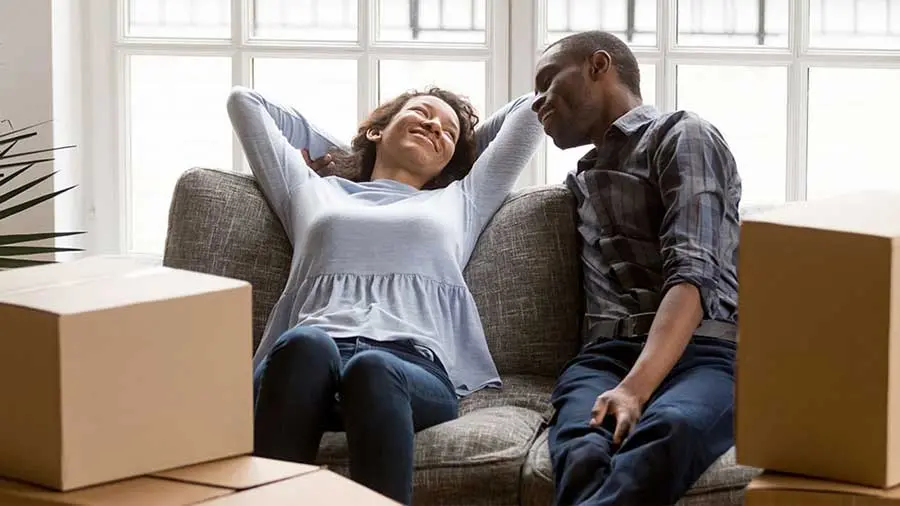 A couple relaxes on a couch surrounded by moving boxes)