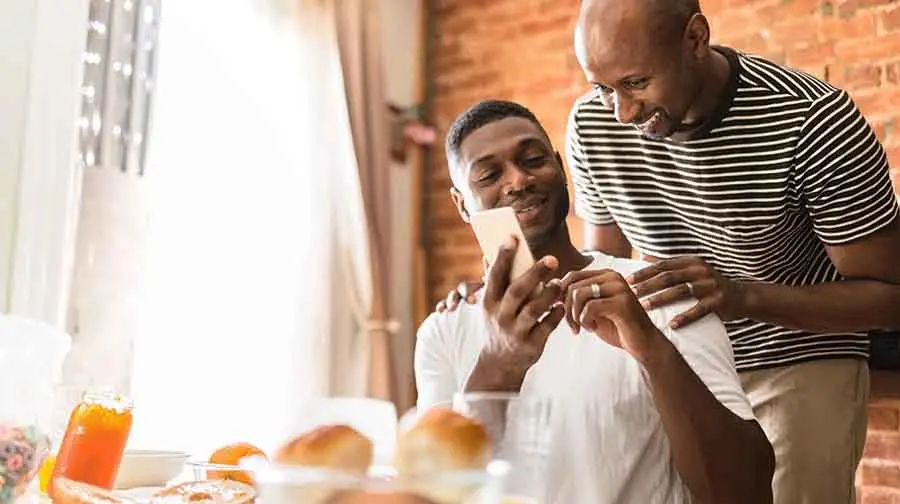 man standing behind partner, both laughing at phone