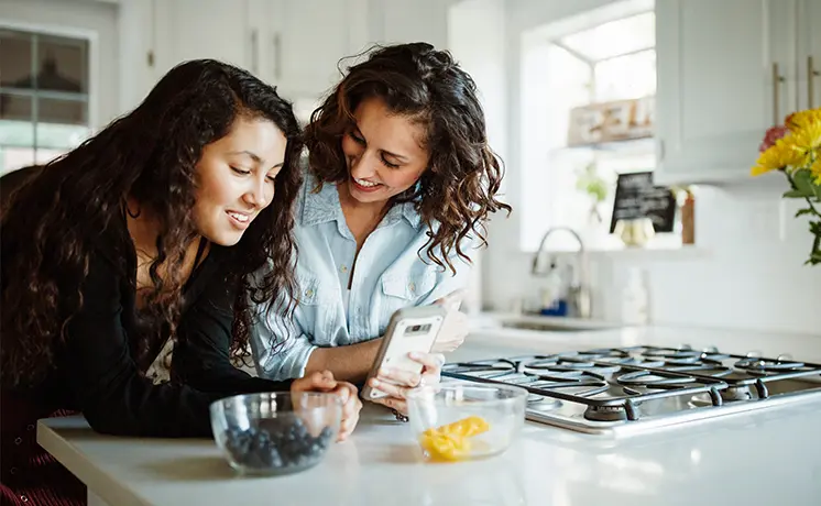 Parent and high school daughter leaning at a kitchen counter to look at something on a phone