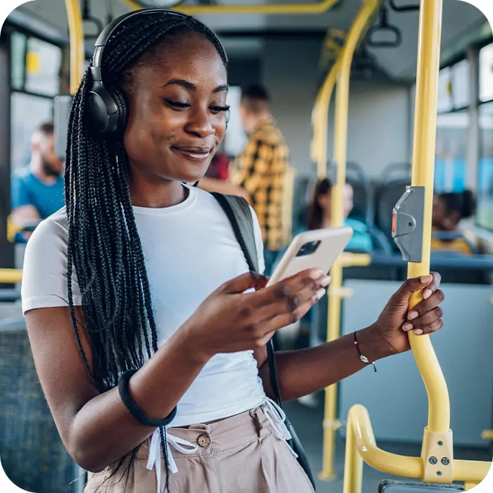 Young adult wearing headphones standing during a train commute and checking her phone in one hand while holding onto pole with her other hand