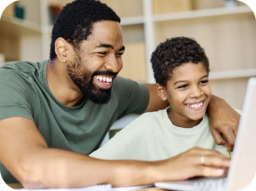 Father with arm around pre-teen while using a laptop, both smiling big