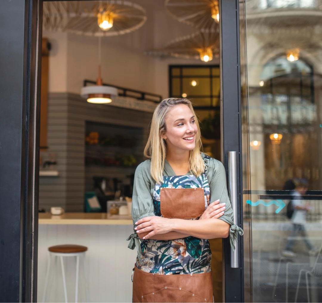 young woman business owner with apron stands outside storefront smiling