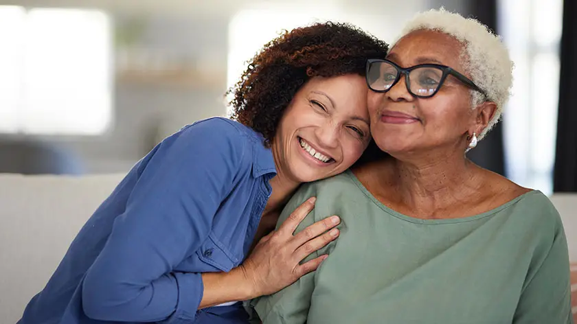 A joyful moment of a daughter warmly hugging her mother, both smiling brightly in a cozy, well-lit home setting, conveying love and connection.