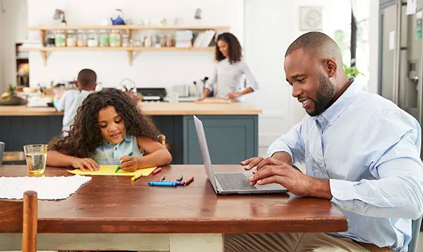 Young falther working on laptop at kitchen table with daughter drawing next to him
