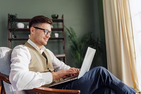 A young man in glasses smiles while working on a laptop.