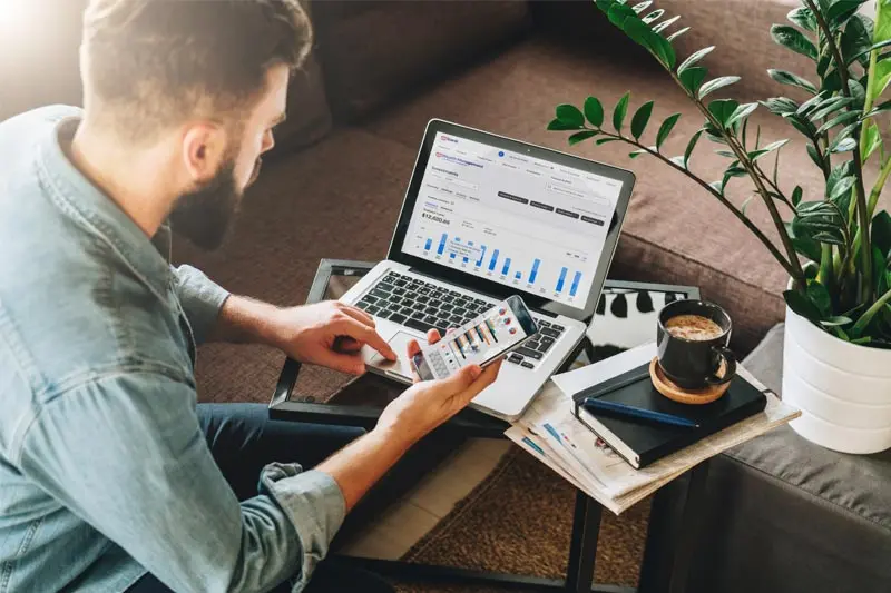 Bearded younger man looking at stock charts on his laptop and phone