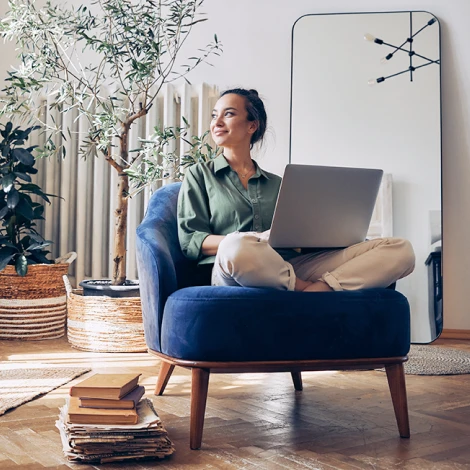 Young woman sitting cross-legged on a chair with a laptop and she is gazing out a window