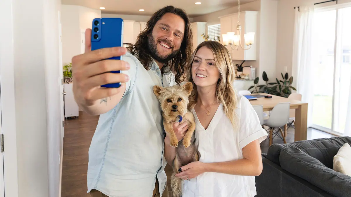 A man and woman take a selfie holding their dog in their new home.