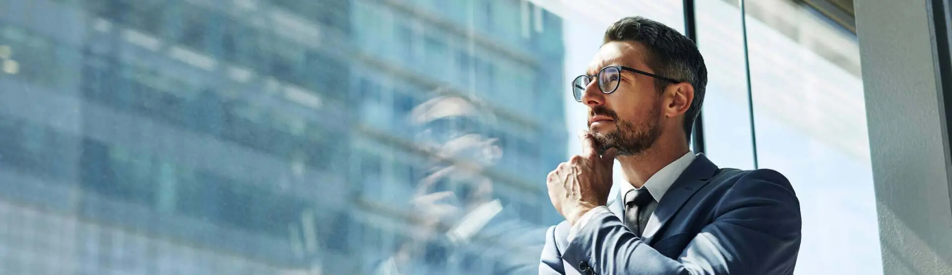 Man wearing suit, arms crossed, gazing out the window of an office 