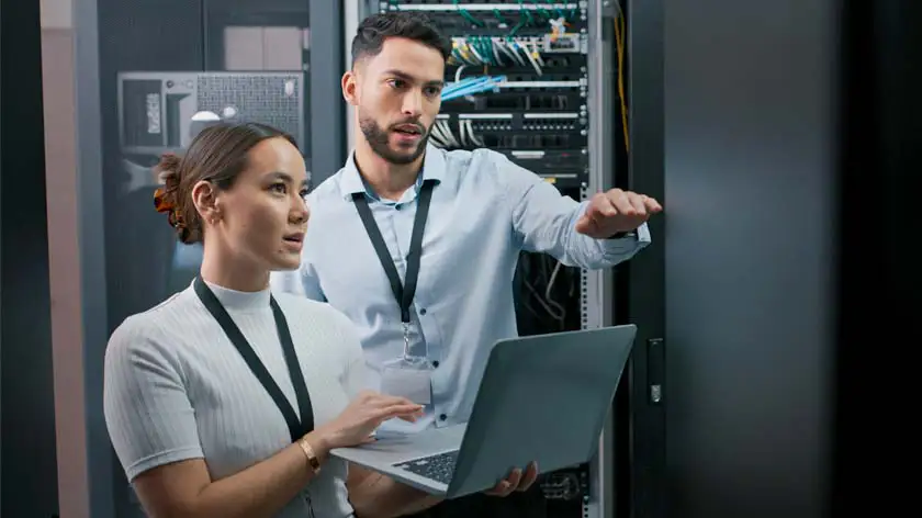 Two IT workers in a server room doing an inspection to help prevent cybersecurity fraud.