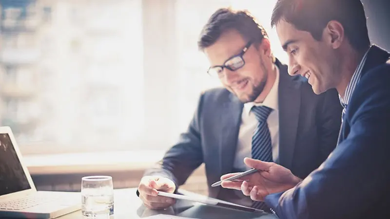 Two men sitting at a desk with a laptop and tablet looking at artificial intelligence and automation trends in investing.
