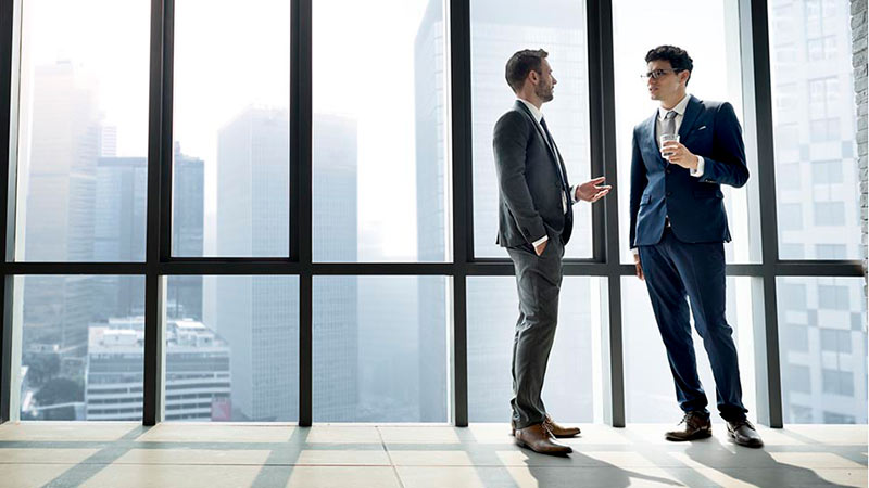 Two colleagues talking in office by large window discussing outsourced chief investment officer trends and benefits. 