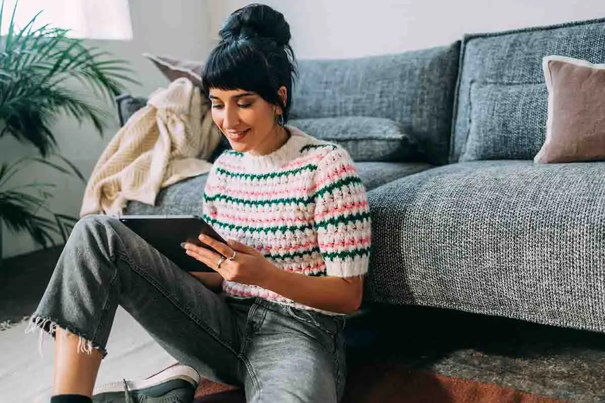 smiling woman working on tablet while leaning against couch in living room