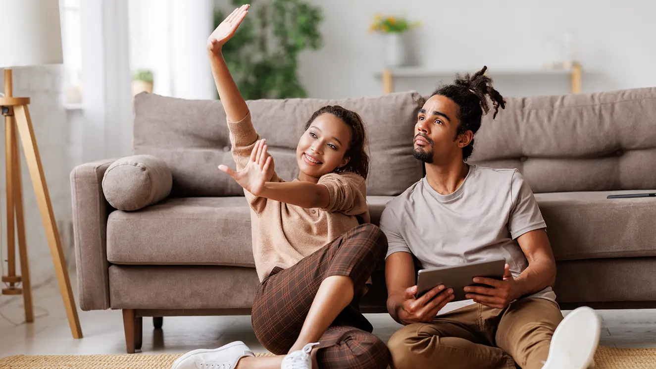 A young woman sitting on the floor with legs crossed, playfully waving hands. A young man next to her looks at a tablet. 