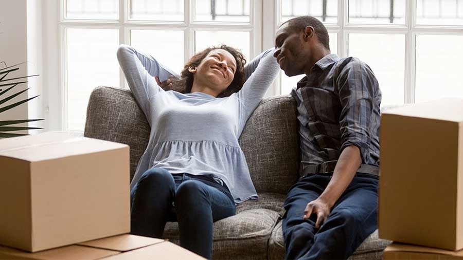 A couple relaxes on a couch surrounded by moving boxes)