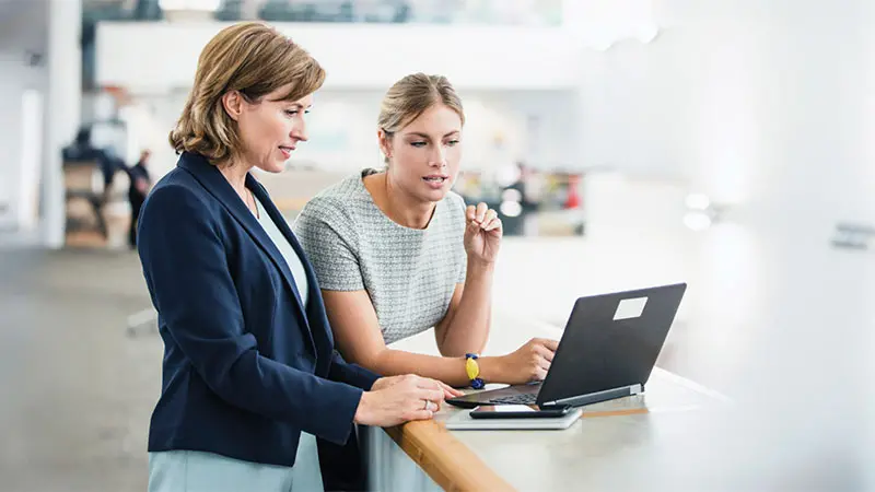 Two professional women standing an looking at a laptop on a desk