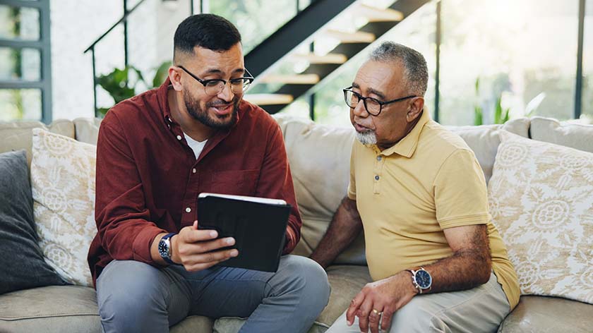 A younger man and his father sitting on a couch, sharing a moment as the son shows something on a tablet, fostering intergenerational connection in a cozy home setting.
