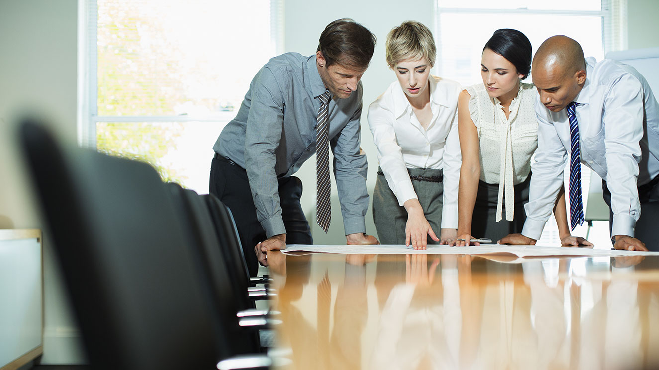 Photo of co-workers, two men and two women, who are standing around the end of a conference table and looking at building plans.