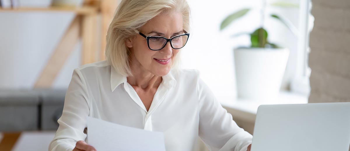 Woman wearing glasses sitting at a desk and holding some paperwork as she reviews her bank account.