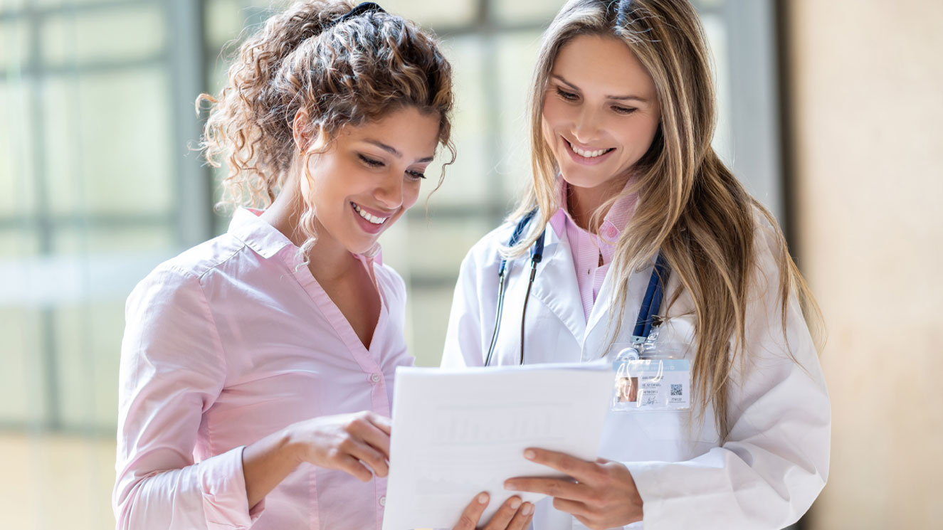 Female doctor and female patient standing next to each other as the doctor is holding a clipboard and explaining paperwork to the patient.