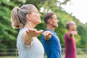Group of senior people with closed eyes stretching arms at park. Happy mature people doing yoga exercise outdoor on a bright morning. Yoga class with woman and men doing breath exercising with stretched arms.